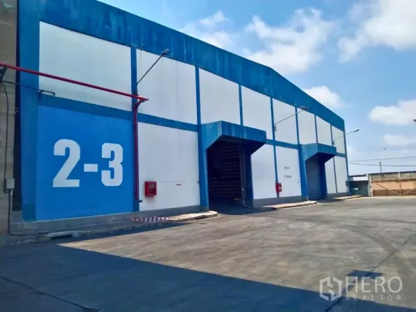 Factory for rent in Samut Prakan - Blue and white factory exterior with roller shutter loading bays and wide truck driveway.