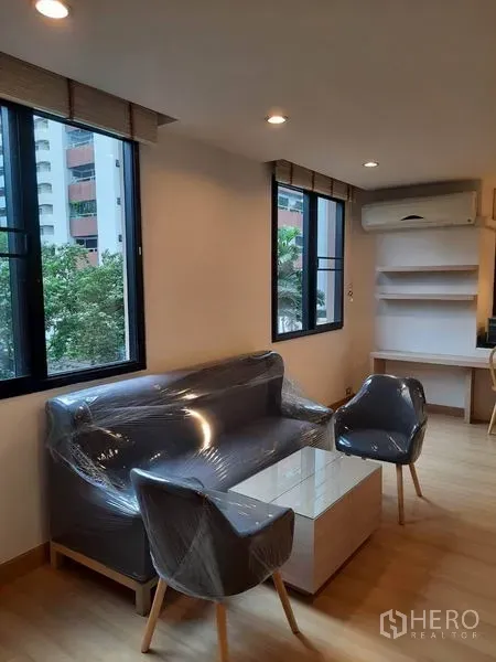 Condominium for rent in Watthana, Bangkok - Bright lounge with large corner windows, compact sofa and armchairs, built-in shelves and air conditioner overlooking greenery.
