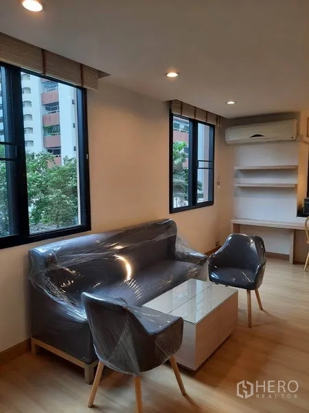 Condominium for rent in Watthana, Bangkok - Bright lounge with large corner windows, compact sofa and armchairs, built-in shelves and air conditioner overlooking greenery.