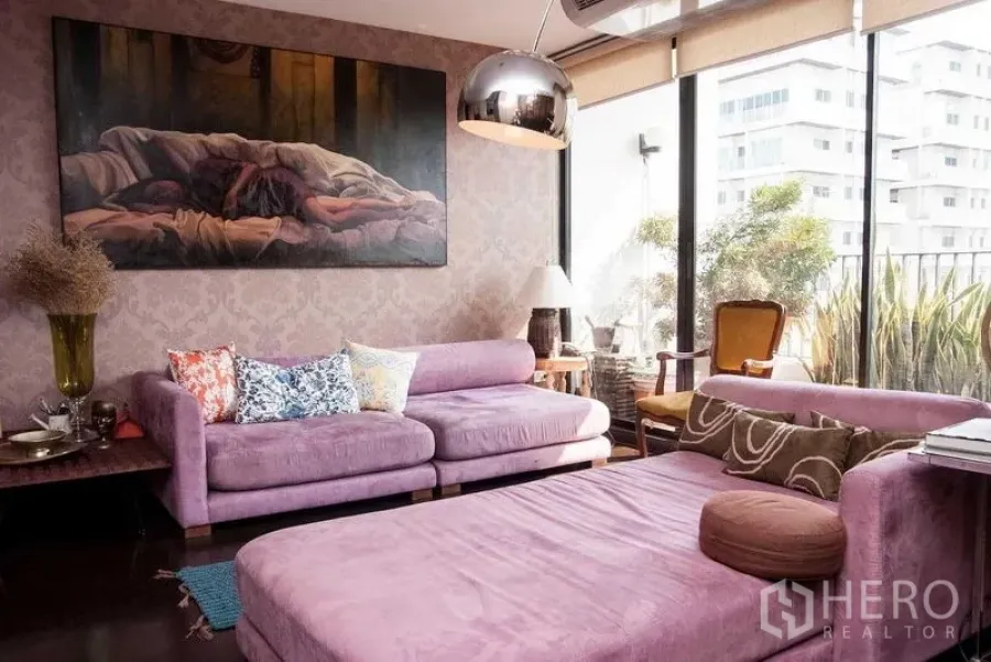 คอนโด ให้เช่าในWatthana, Bangkok - ห้องนั่งเล่นโซฟาสีม่วง หน้าต่างเต็มบานสู่ระเบียงมีต้นไม้ โคมไฟโดมโลหะ และภาพศิลป์ขนาดใหญ่