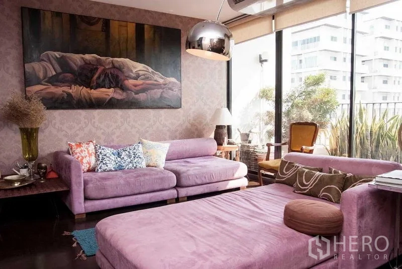 คอนโด ให้เช่าในWatthana, Bangkok - ห้องนั่งเล่นโซฟาสีม่วง หน้าต่างเต็มบานสู่ระเบียงมีต้นไม้ โคมไฟโดมโลหะ และภาพศิลป์ขนาดใหญ่