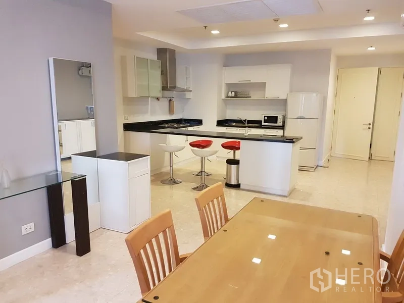 Condominium for rent in Khlong Toei, Bangkok - Open-plan kitchen with white cabinetry, black stone countertops, island breakfast bar with red stools, and adjacent dining area.