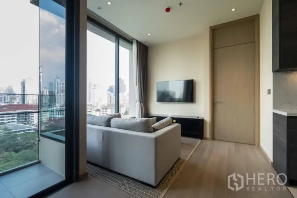 公寓 出租在Watthana, Bangkok - 明亮客厅，沙发、挂墙电视与落地窗，连接阳台可眺望城市天际线。