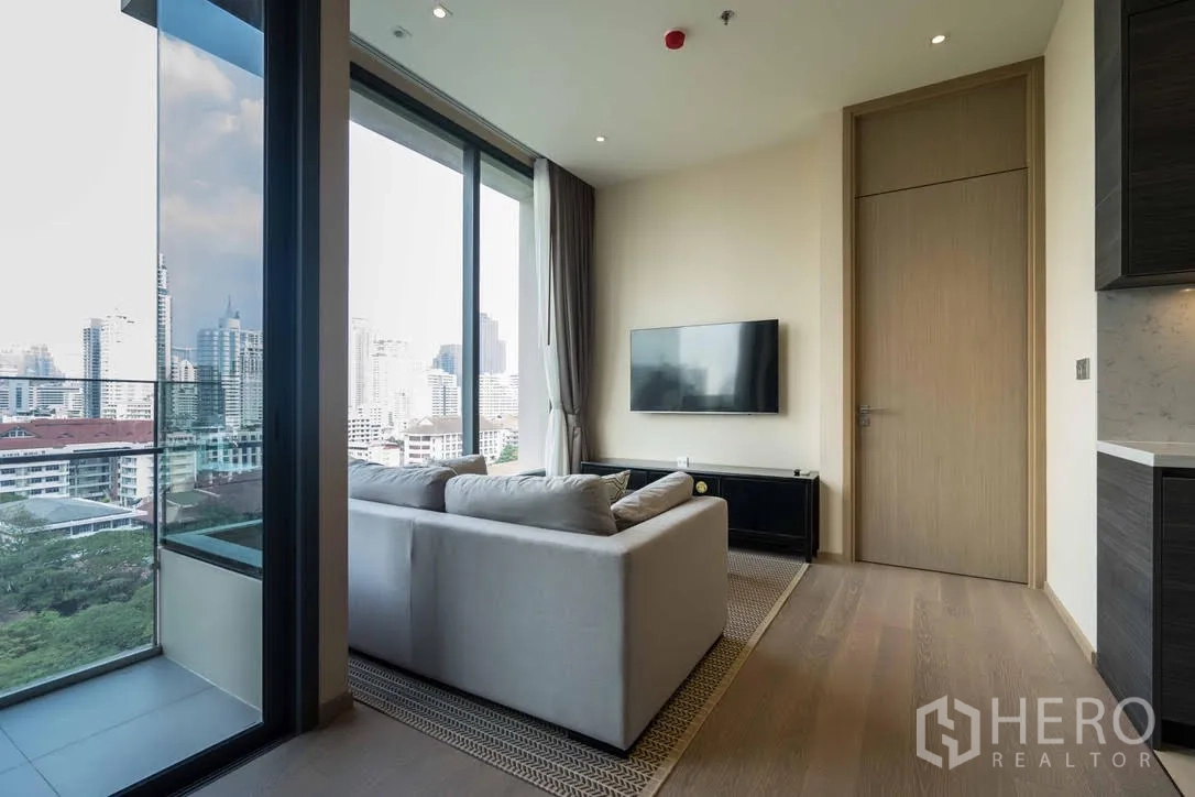 公寓 出租在Watthana, Bangkok - 明亮客厅，沙发、挂墙电视与落地窗，连接阳台可眺望城市天际线。