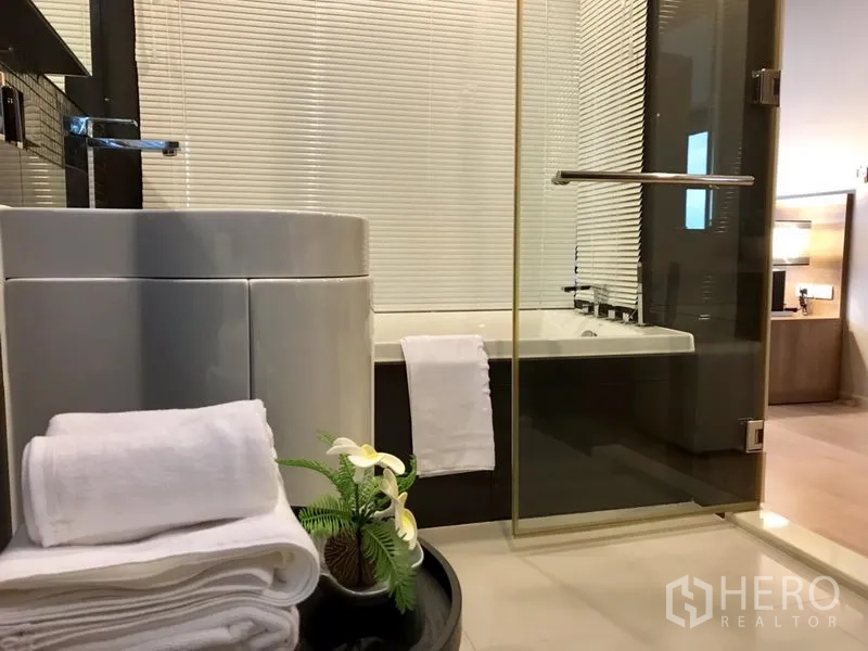 Condominium for rent in Khlong Toei, Bangkok - Bathroom with glass screen, bathtub, round vanity and fresh white towels.