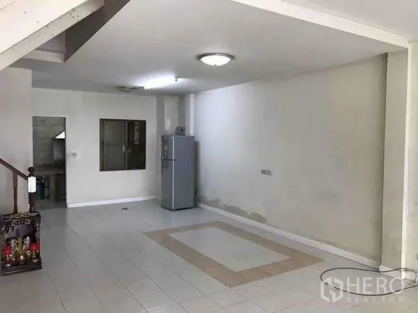 Townhouse for rent in Suan Luang, Bangkok - Open-plan living area with white tiled floor, ceiling lights and a refrigerator leading toward the kitchen.