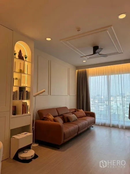 Condominium for rent in Bang Rak, Bangkok - Bright lounge with floor-to-ceiling curtains, brown sofa, built-in bookcase and glass doors to the balcony.