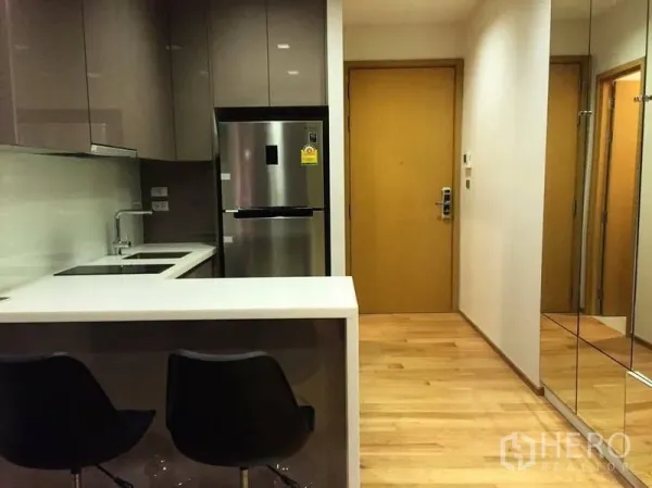 Condominium for rent or sale in Watthana, Bangkok - Compact modern kitchen with breakfast counter, two bar stools, mirrored wall and wooden entry door.