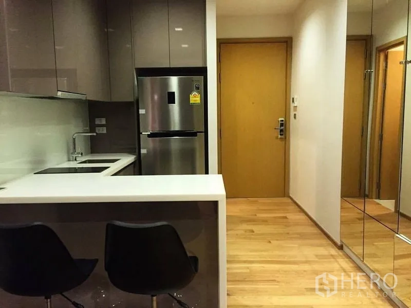 Condominium for rent or sale in Watthana, Bangkok - Compact modern kitchen with breakfast counter, two bar stools, mirrored wall and wooden entry door.