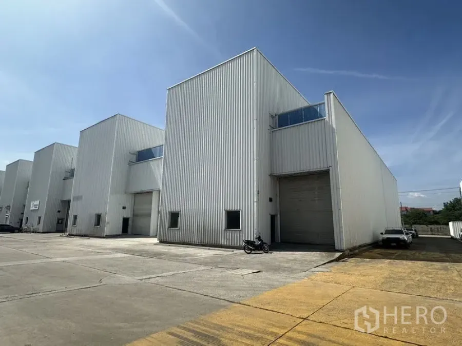 Factory for rent in Khlong Luang, Pathum Thani - Row of modern warehouse units with high roller doors and parking area in an industrial compound.