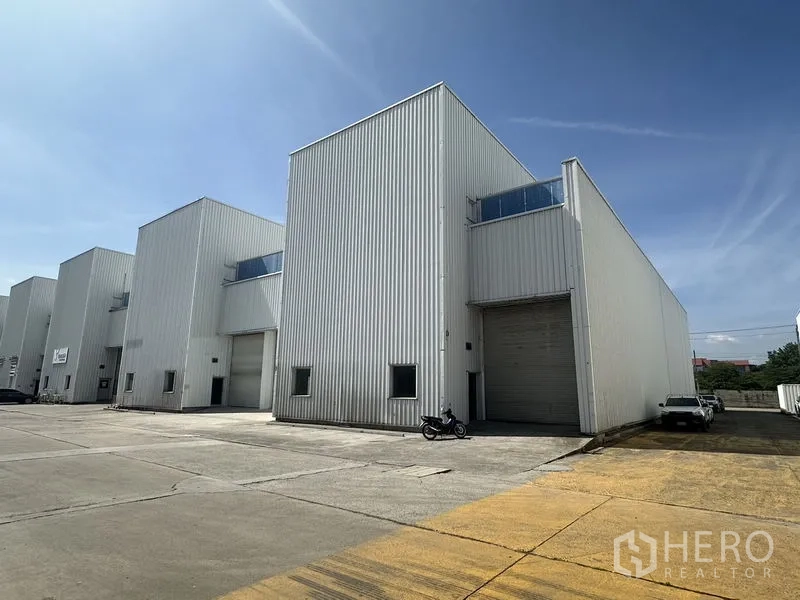 Factory for rent in Khlong Luang, Pathum Thani - Row of modern warehouse units with high roller doors and parking area in an industrial compound.