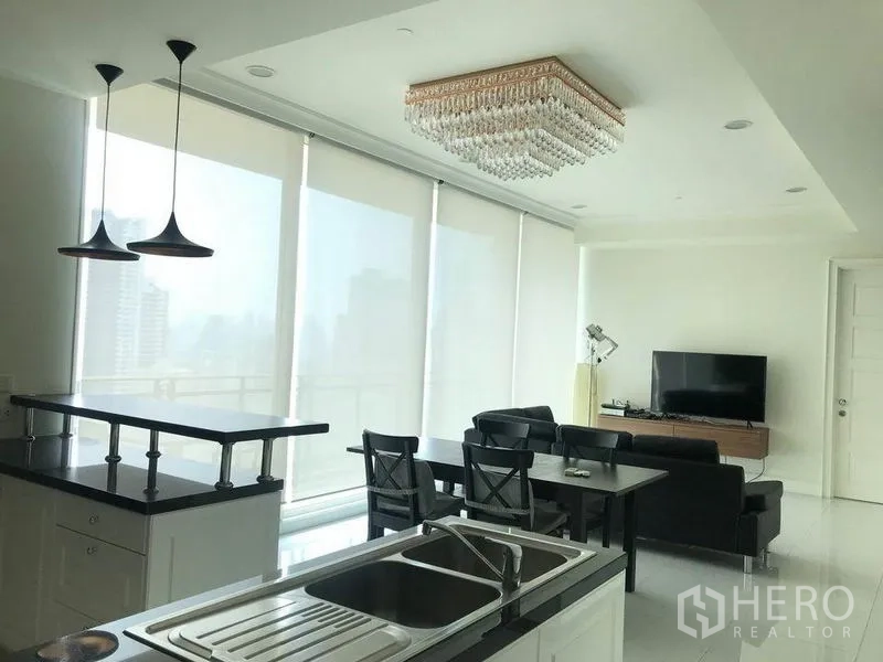 Condominium for rent in Watthana, Bangkok - Kitchen island with double sink facing dining table and TV lounge under a crystal chandelier.