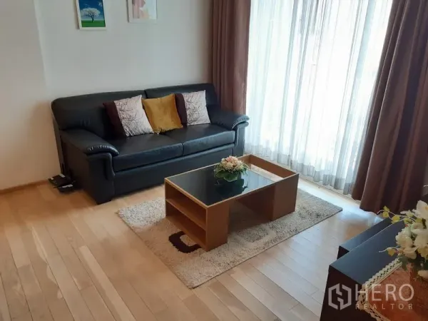 Condominium for rent in Khlong Toei, Bangkok - Bright living room with a black leather sofa, coffee table, and floor-to-ceiling curtains letting in natural light.