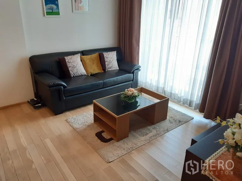 Condominium for rent in Khlong Toei, Bangkok - Bright living room with a black leather sofa, coffee table, and floor-to-ceiling curtains letting in natural light.
