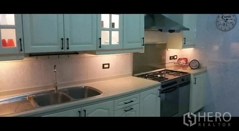 Condominium for rent in Khlong Toei, Bangkok - Modern kitchen with white cabinets, double sink, gas stove and range hood under warm task lighting.