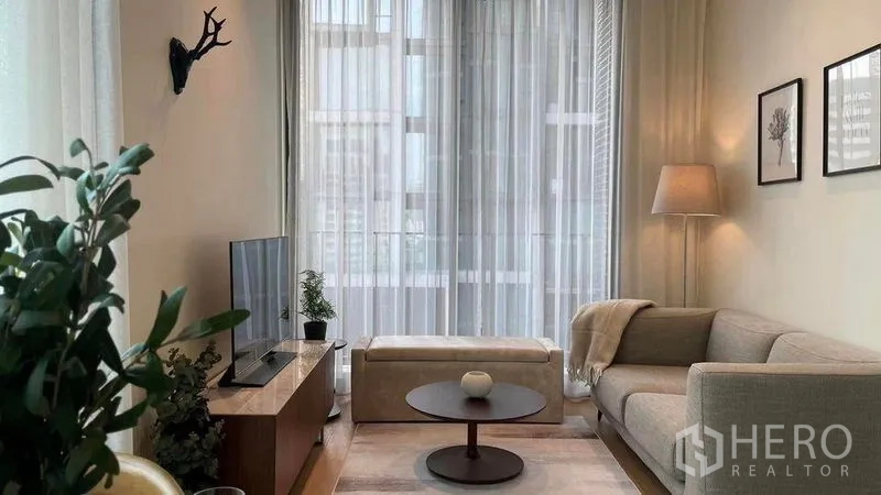 Condominium for rent in Pathum Wan, Bangkok - Bright living room with sofa, round coffee table, TV console and sheer curtains facing city buildings.