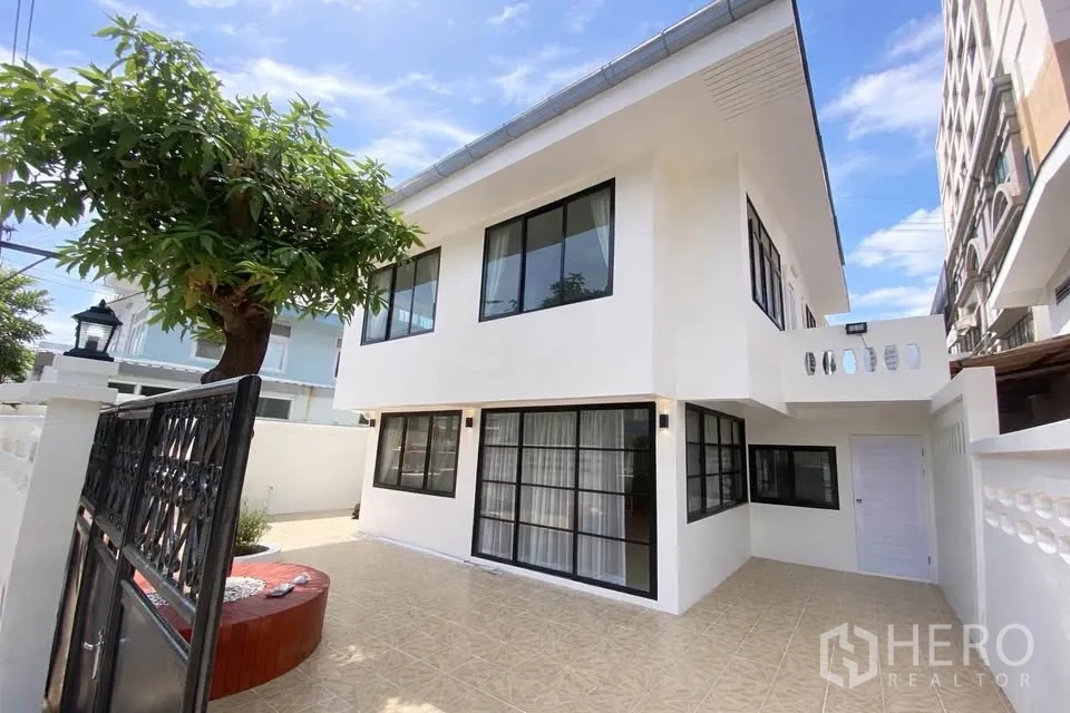 บ้านเดี่ยว for rent in Huai Khwang, Bangkok - ภายนอกบ้านเดี่ยวสองชั้นสีขาว มีลานกระเบื้องและต้นไม้ภายในรั้ว