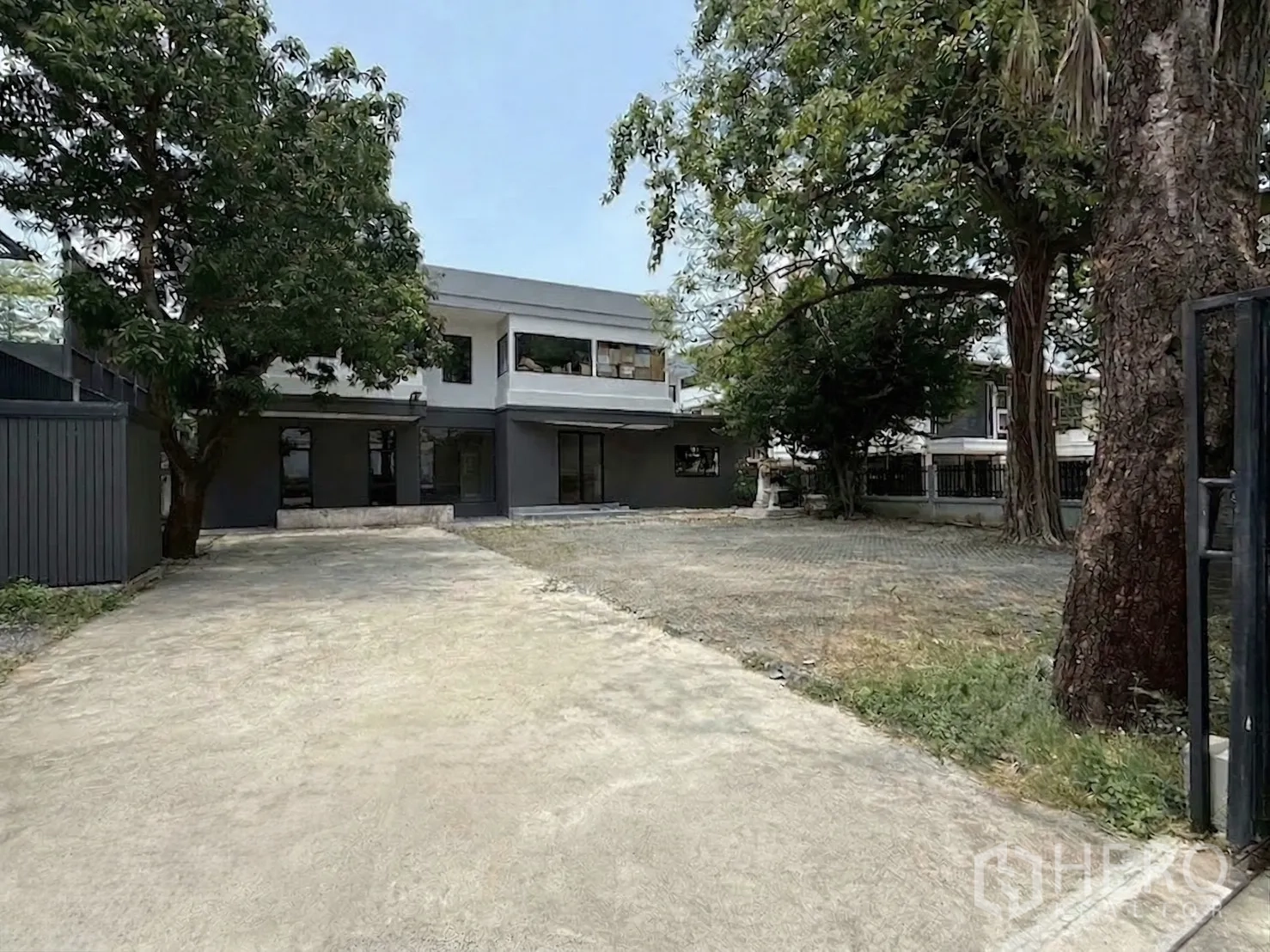 Home Office for rent in Watthana, Bangkok - Gated driveway leading to a two-storey modern home office amid mature trees.