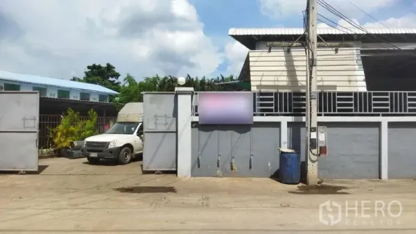 Warehouse for sale in Samut Sakhon - Front facade of the warehouse showing secure wall, double metal gate and roadside utility pole.