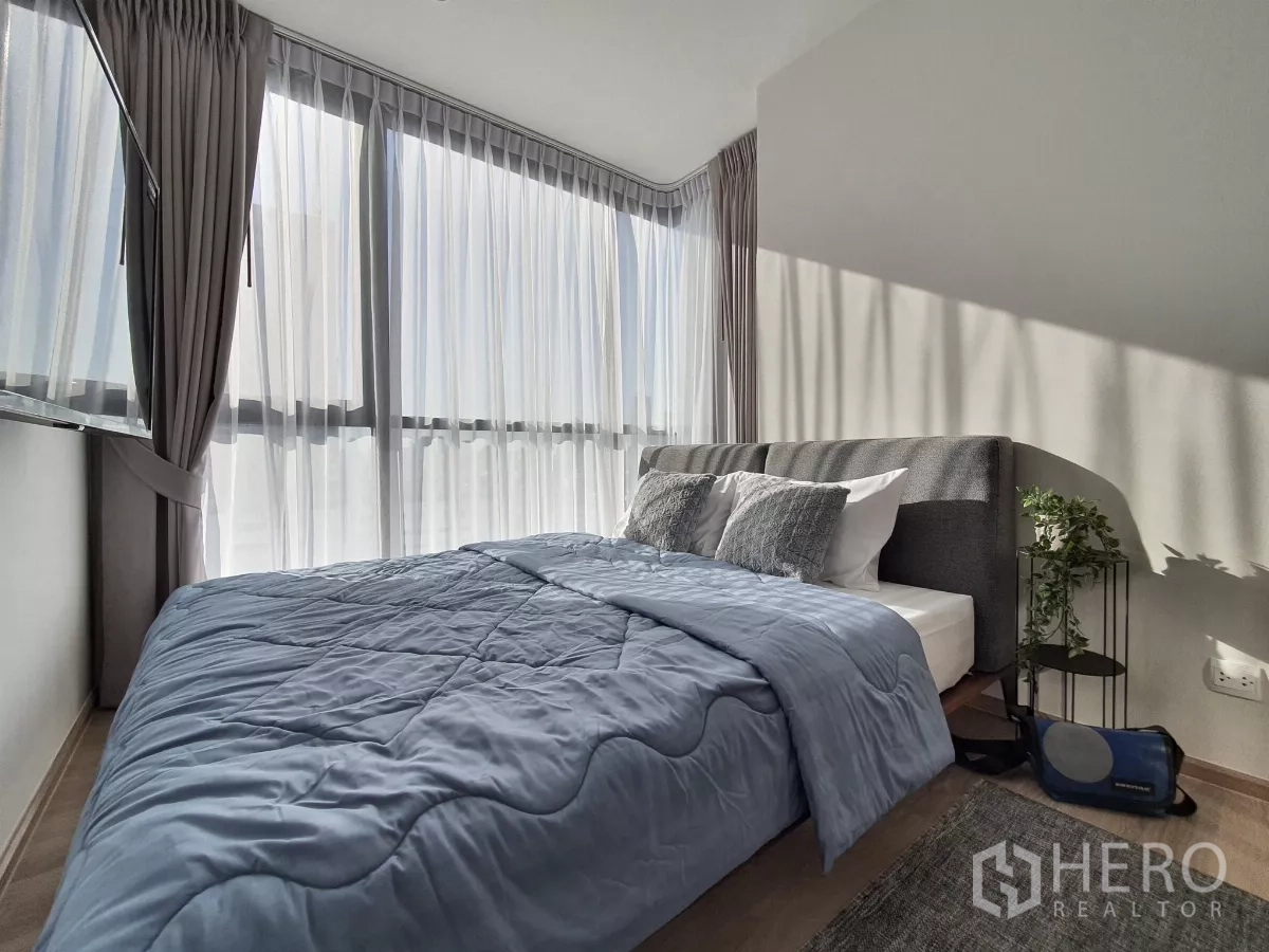 Condominium for rent in Khlong Toei, Bangkok - Corner bedroom with sheer curtains, city light and bedside plant.
