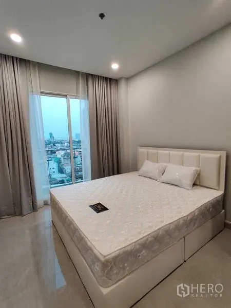 คอนโด ให้เช่าในBang Rak, Bangkok - ห้องนอนเตียงบุนวม หน้าต่างบานใหญ่เห็นวิวเมือง ผ้าม่านสีเทา และไฟดาวน์ไลท์