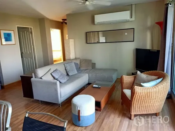 Condominium for rent in Khlong Toei, Bangkok - Cozy living area with sectional sofa, rattan chair, TV and a long wall mirror.