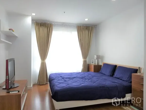 Condominium for rent or sale in Khlong Toei, Bangkok - Sunlit bedroom with blue bedding on a double bed, bedside drawers and floor-to-ceiling curtains.