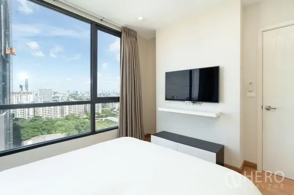 公寓 出租在Watthana, Bangkok - 卧室设落地窗与开阔城市景观，墙面安装电视，简约配色。