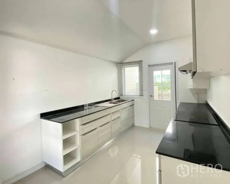 Detached House for sale in Bang Khen, Bangkok - Bright kitchen with long black stone countertops, white built-in cabinets, window and back door.