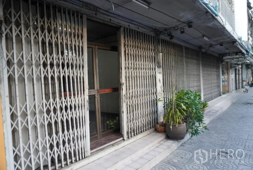 Shophouse for rent in Khlong Toei, Bangkok - Close-up of a shop entrance with folding steel doors, glass inner door, and potted plants along the pavement.