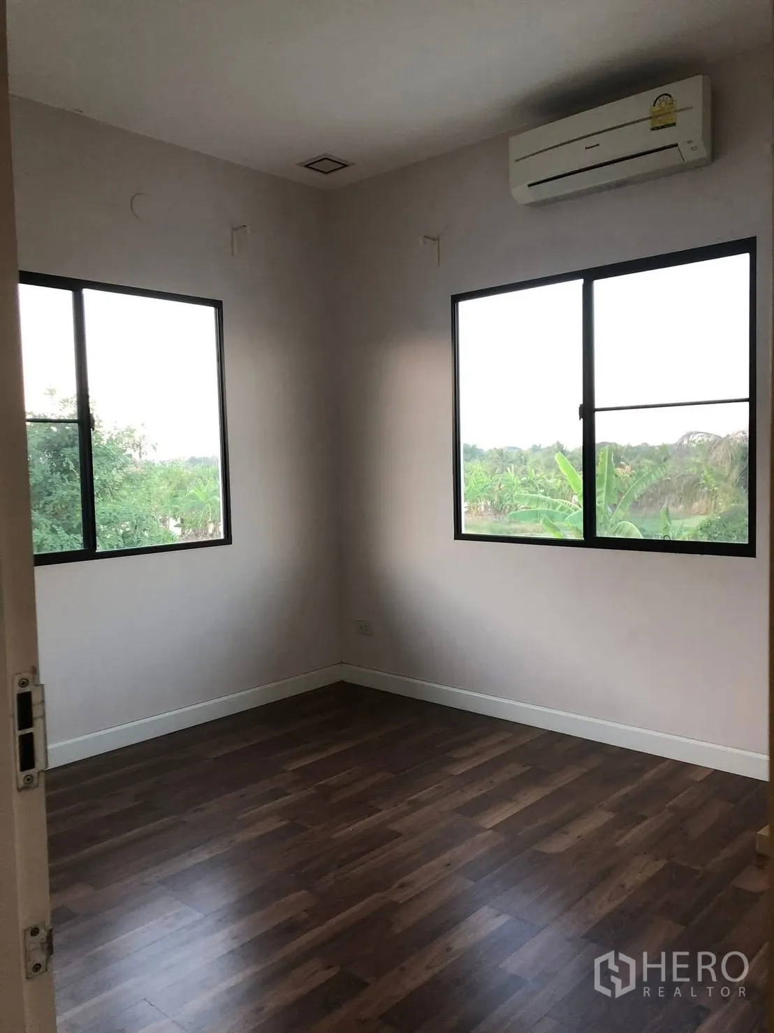 Detached House for rent in Prawet, Bangkok - Corner bedroom with wood flooring, two windows and a wall-mounted air conditioner overlooking greenery.