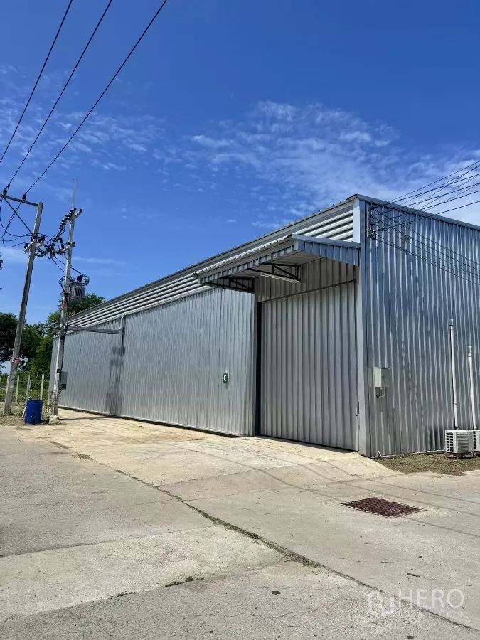 Warehouse for rent in Bang Lamung, Chon Buri - Modern metal-clad warehouse exterior with wide sliding door, concrete apron and power lines under a clear blue sky.