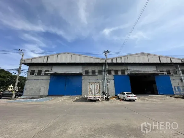 คลังสินค้า ให้เช่าในBang Phli, Samut Prakan - มุมมองด้านหน้าที่มีประตูเหล็กสีน้ำเงินหลายบานและที่จอดรถหน้าโกดัง