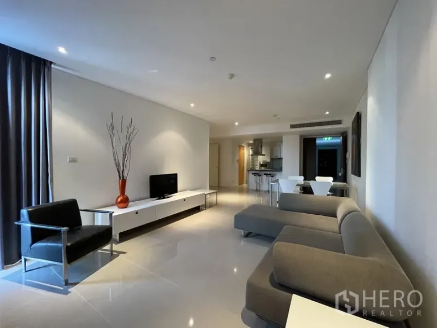 Condominium for rent or sale in Watthana, Bangkok - Open-plan living room with gray L-shaped sofa, TV console, black armchair, and floor-to-ceiling curtains leading toward the kitchen.