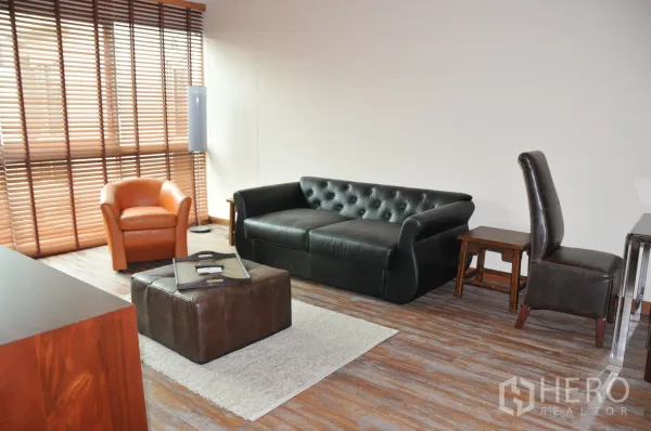 Condominium for rent in Khlong Toei, Bangkok - Bright living room with black leather sofa, orange armchair, ottoman, and wooden blinds.