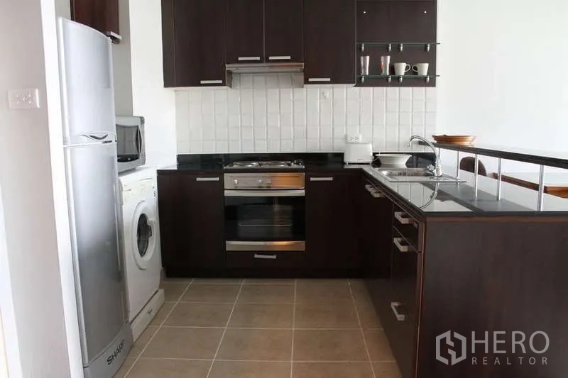 Condominium for rent in Sathon, Bangkok - Full view of L-shaped kitchen featuring fridge, washing machine, oven, and black stone countertops.