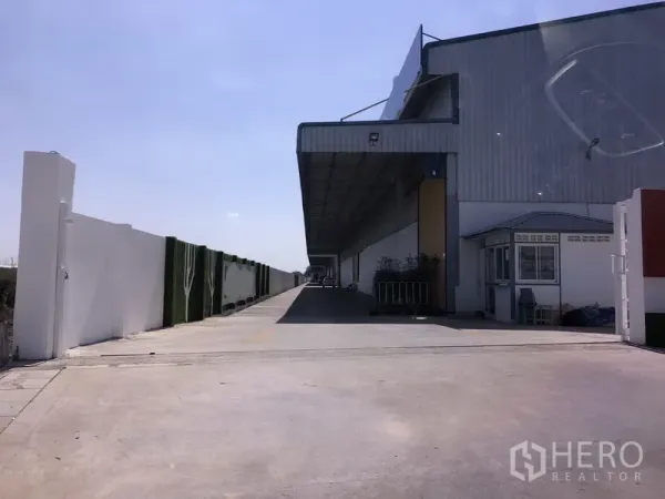 货栈 出租在Lat Krabang, Bangkok - 曼谷Lat Krabang仓库外观，带长型遮雨装卸平台、宽阔混凝土车道、围墙与门卫岗亭，天空晴朗。