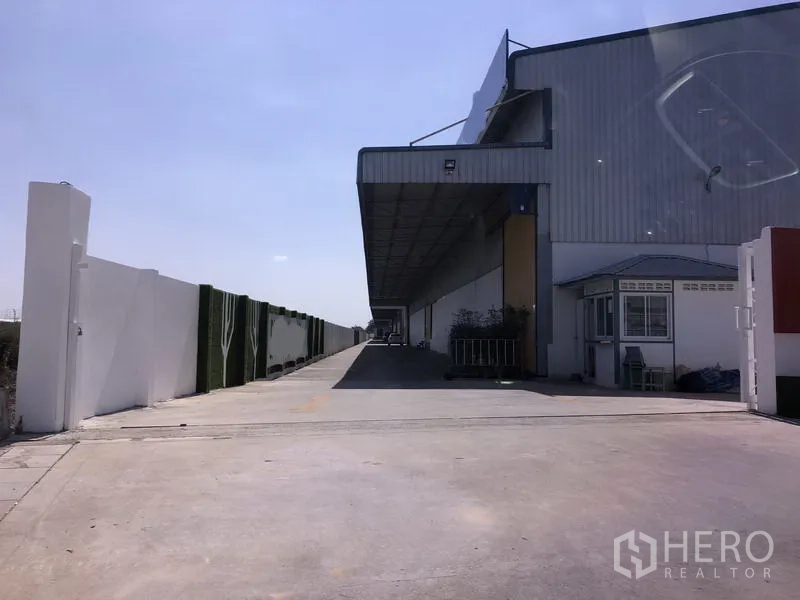 货栈 出租在Lat Krabang, Bangkok - 曼谷Lat Krabang仓库外观，带长型遮雨装卸平台、宽阔混凝土车道、围墙与门卫岗亭，天空晴朗。