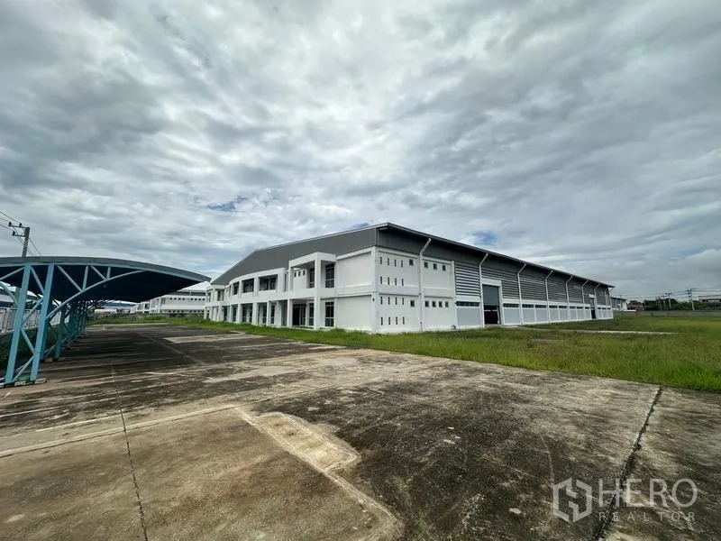 Factory for rent in Pathum Thani - Modern factory exterior with wide driveway and covered parking under cloudy sky.