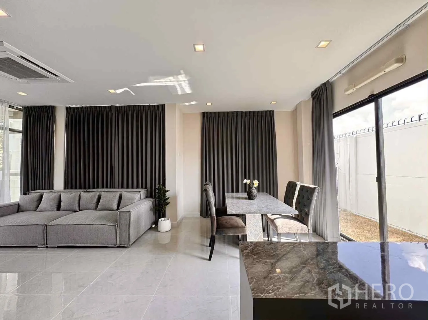 บ้านเดี่ยว for rent in Bang Phli, Samut Prakan - พื้นที่นั่งเล่น-ทานอาหารพร้อมโซฟาสีเทา ชุดทานข้าว และประตูกระจกบานเลื่อนออกสู่สนาม.