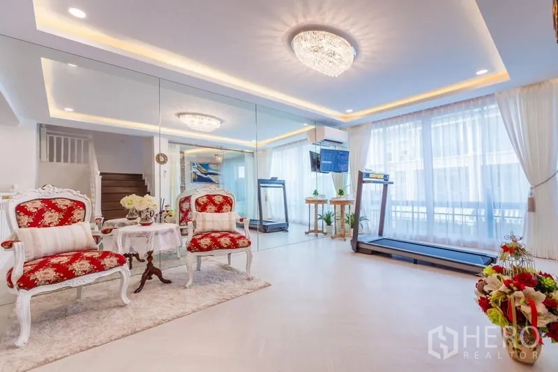 Townhouse for rent in Lat Phrao, Bangkok - Bright living area with mirrored wall, chandelier lighting, classic red armchairs and a treadmill by floor‑to‑ceiling curtains.