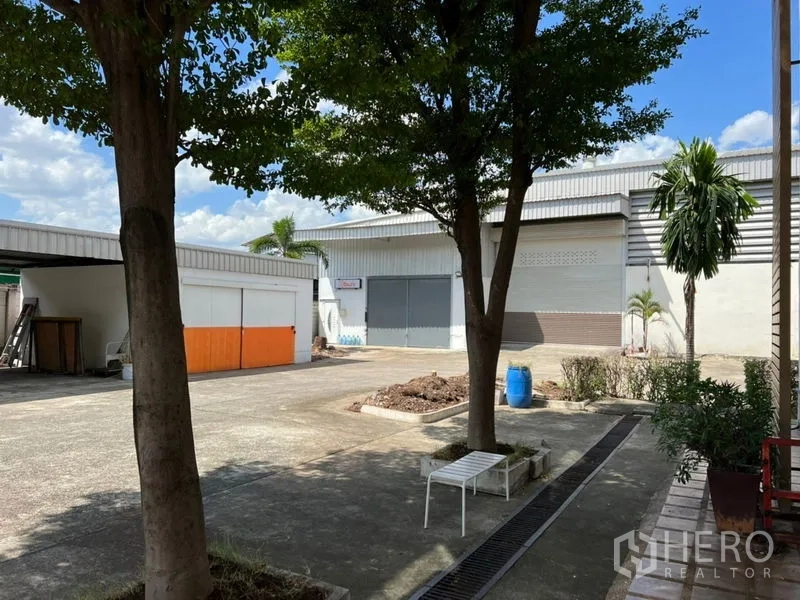 Warehouse for rent in Prawet, Bangkok - Open courtyard with shade trees, drainage channel and surrounding warehouse buildings with roller doors and utility structures.
