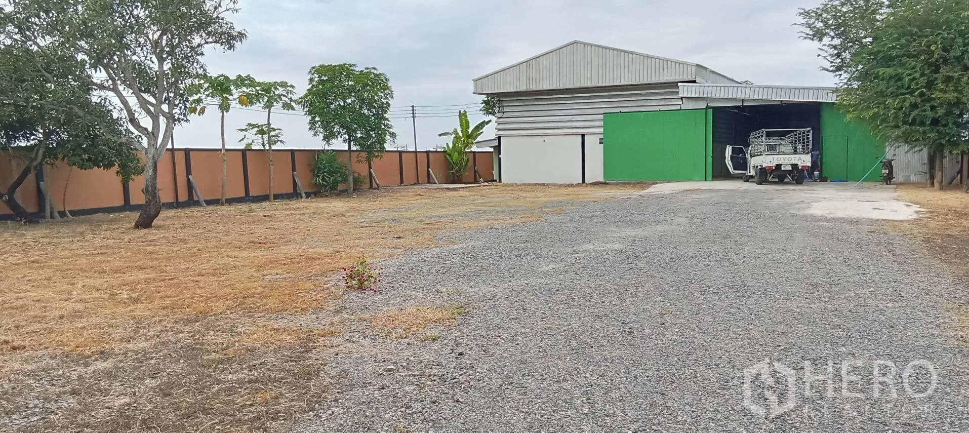 โกดัง for sale in Lam Luk Ka, Pathum Thani - พื้นที่โกดังมีกำแพงรั้ว ประตูทางเข้า ลานหินคลุกกว้าง และทางรถขนถ่ายสินค้า