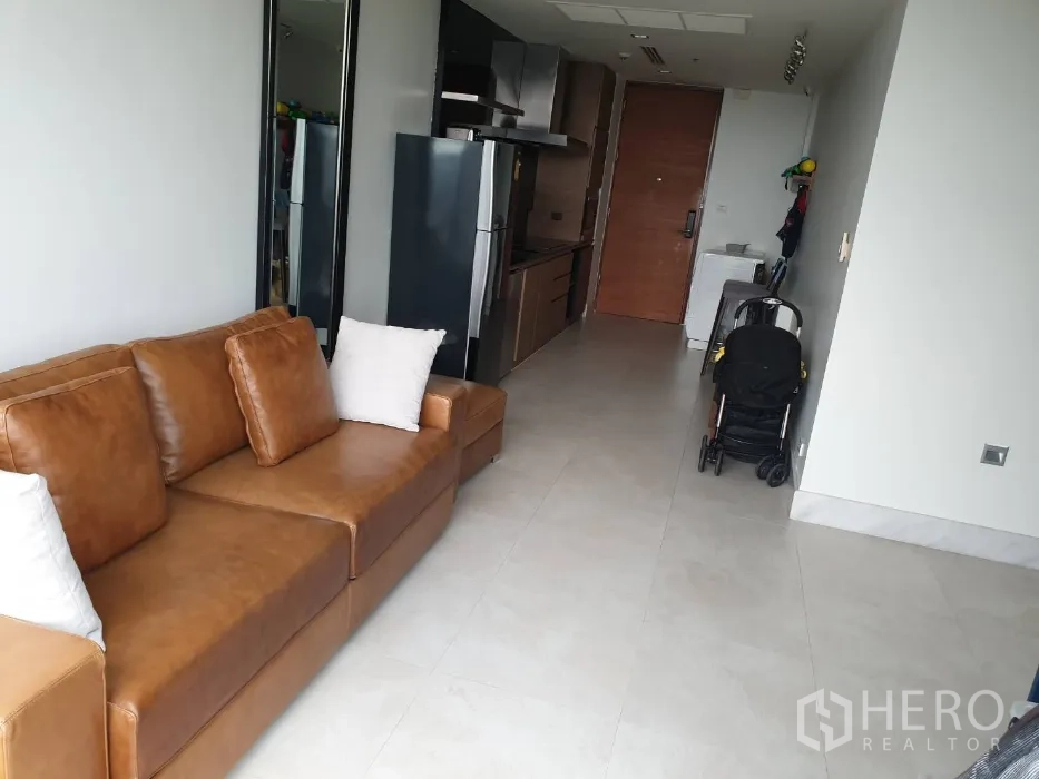 Condominium for rent in Khlong Toei, Bangkok - Open-plan living room with a brown leather sofa, tiled floors, and a compact corridor kitchen in a Bangkok condo.
