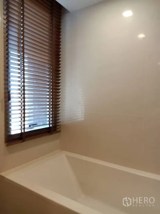 Condominium for rent in Khlong Toei, Bangkok - Modern bathroom with built-in bathtub beside a window fitted with wooden blinds.