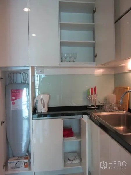 Condominium for rent in Khlong Toei, Bangkok - Compact kitchen with glossy white cabinets, glass backsplash, sink and ample storage.