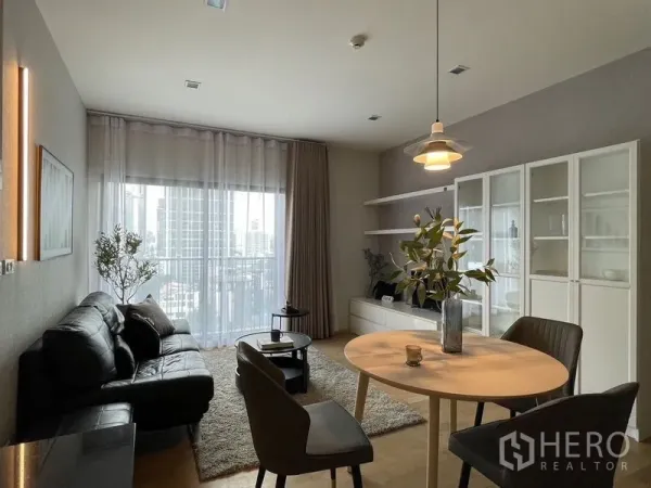 Condominium for rent in Watthana, Bangkok - Open-plan living and dining area with black leather sofa, round table, and floor-to-ceiling window showing a city view.
