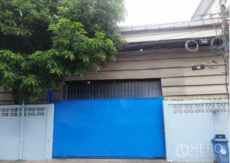 คลังสินค้า ให้เช่าในBang Kapi, Bangkok - ประตูรั้วสีฟ้าและด้านหน้าโกดังชั้นเดียว มีต้นไม้ให้ร่มเงา