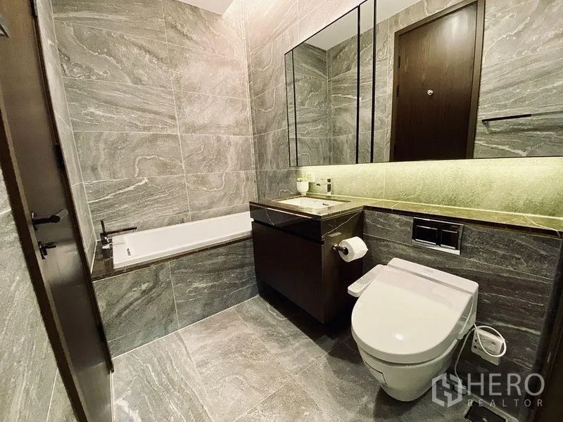 Condominium for rent in Khlong Toei, Bangkok - Hotel-style bathroom with gray stone tiles, vanity mirror cabinet, bathtub, and smart toilet.