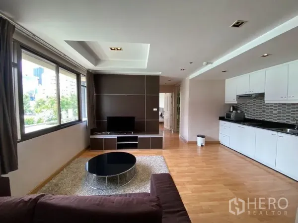 Condominium for rent in Khlong Toei, Bangkok - Bright lounge with TV wall, tray ceiling, and city-facing windows beside a kitchenette.